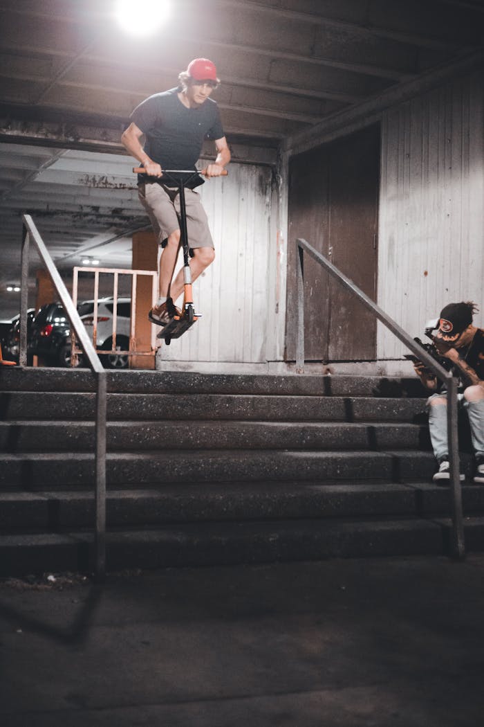 A young man performs a scooter jump down city stairs at night with a friend capturing the moment.