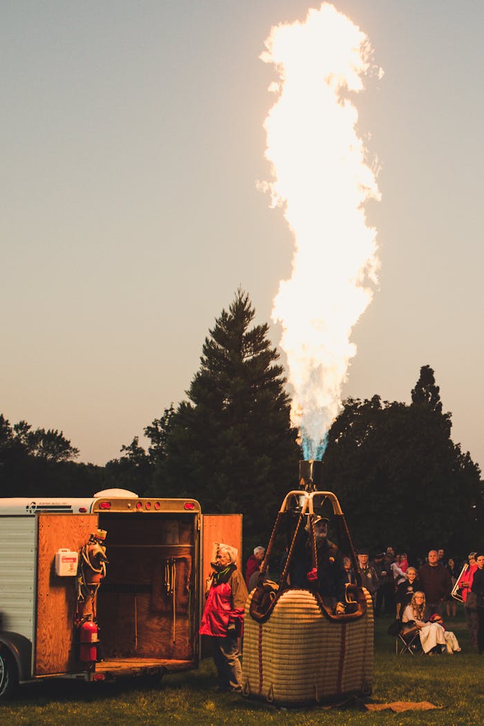 A group prepares a hot air balloon burner for flight at twilight, surrounded by trees and spectators.